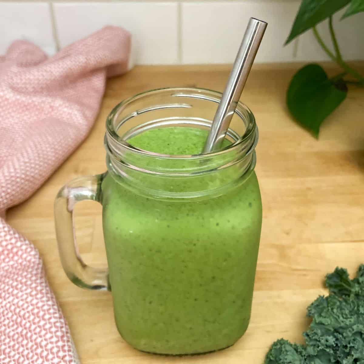 Green kale protein smoothie served in a glass mason jar with a metal straw, placed on a wooden surface next to fresh kale leaves and a pink textured cloth.