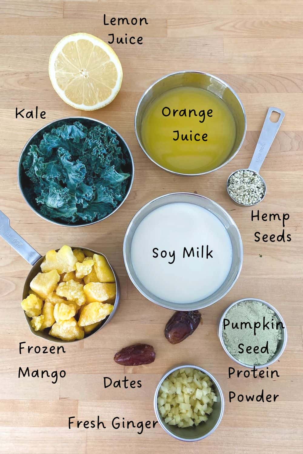  Flat lay of kale protein smoothie ingredients on a wooden surface, including frozen mango, kale, soy milk, orange juice, lemon juice, dates, fresh ginger, hemp seeds, and pumpkin seed protein powder, each labeled with text.