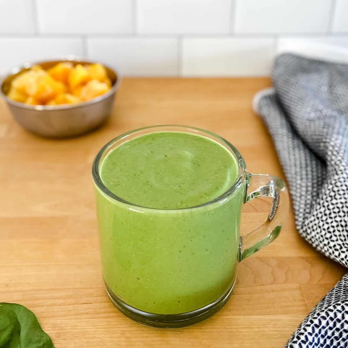 Creamy spinach protein smoothie in a glass cup with handle on wooden counter, black and white tea towel, and bowl of frozen mango in background.