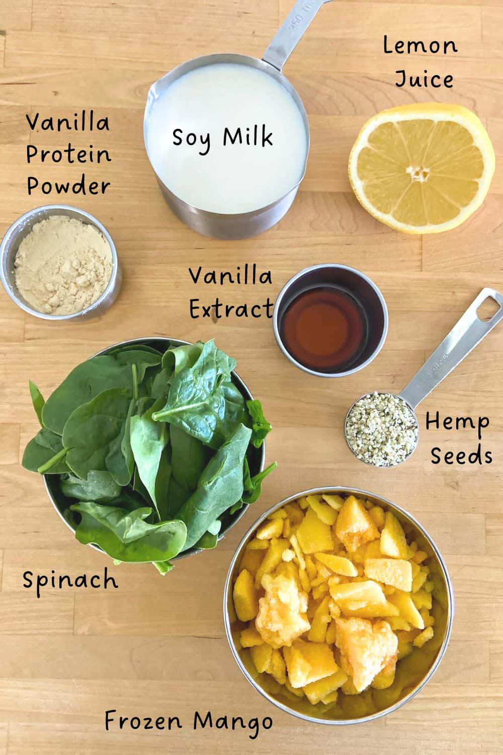 Top-down view of spinach protein smoothie ingredients: spinach, frozen mango, protein powder, soy milk, lemon juice, vanilla extract, hemp seeds.