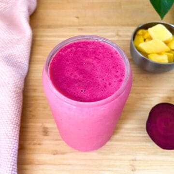 Bright pink smoothie in a clear glass on a wooden surface, with a small bowl of frozen mango chunks and a sliced raw beet in the background.