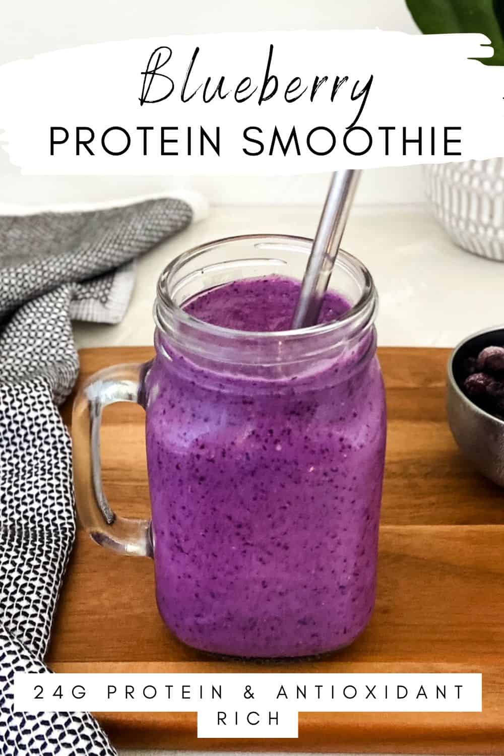 Blueberry protein smoothie in a glass jar with a stainless steel straw on a wooden surface with a linen cloth and a bowl of blueberries in the background.