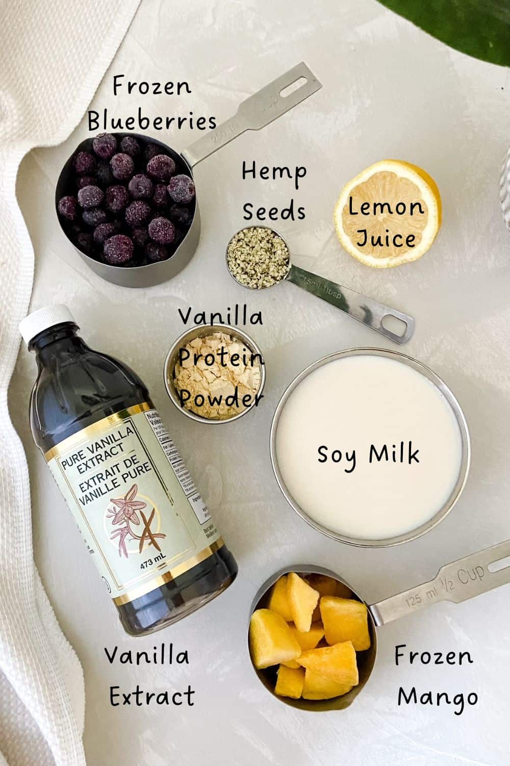 Overhead photo of smoothie ingredients including frozen blueberries, mango, soy milk, vanilla extract, hemp seeds, lemon juice, and vanilla protein powder in measuring cups.