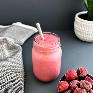 Pink cherry smoothie served in a mason jar with a metal straw, placed on a dark surface with a bowl of frozen cherries and a textured cloth nearby.