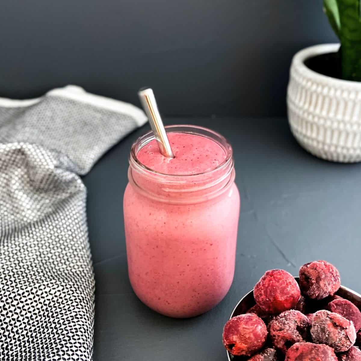 Pink cherry smoothie served in a mason jar with a metal straw, placed on a dark surface with a bowl of frozen cherries and a textured cloth nearby.