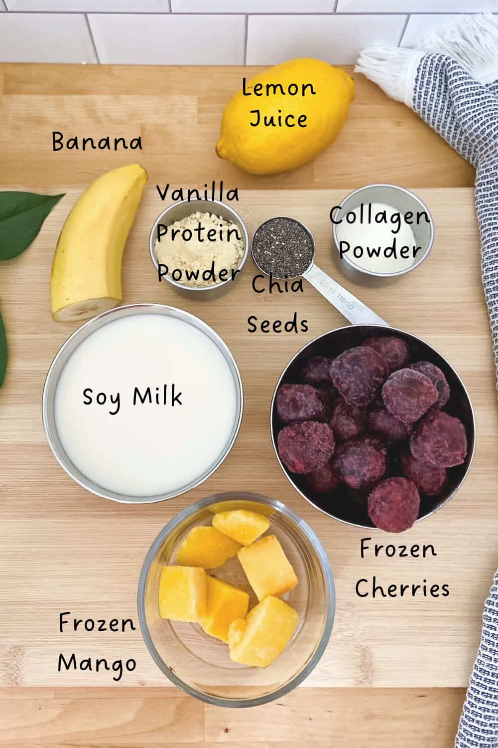 Flat lay of cherry protein smoothie ingredients on a wooden surface, including soy milk, frozen cherries, frozen mango, banana, lemon, chia seeds, collagen powder, and vanilla protein powder, each labeled in black text.
