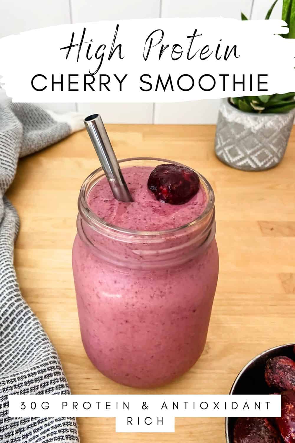 Cherry smoothie in a mason jar topped with a cherry and a stainless steel straw, styled on a wooden surface with a cloth and bowl of frozen cherries nearby. Text overlay reads High Protein Cherry Smoothie and 30g Protein & Antioxidant Rich.