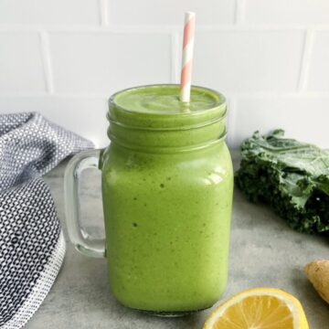 Green kale protein smoothie in a glass mason jar with a striped paper straw on a grey countertop, with fresh kale, ginger, and lemon nearby.