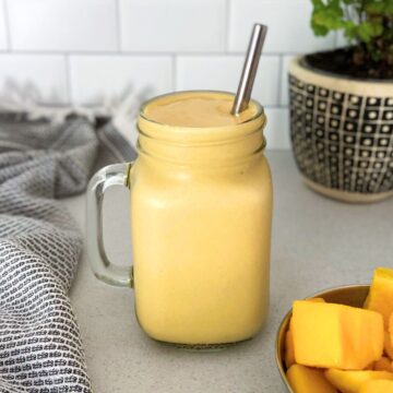 Glass jar filled with a thick and creamy mango protein smoothie, served with a stainless steel straw, a bowl of fresh mango chunks, a patterned cloth napkin, and a decorative plant in the background.