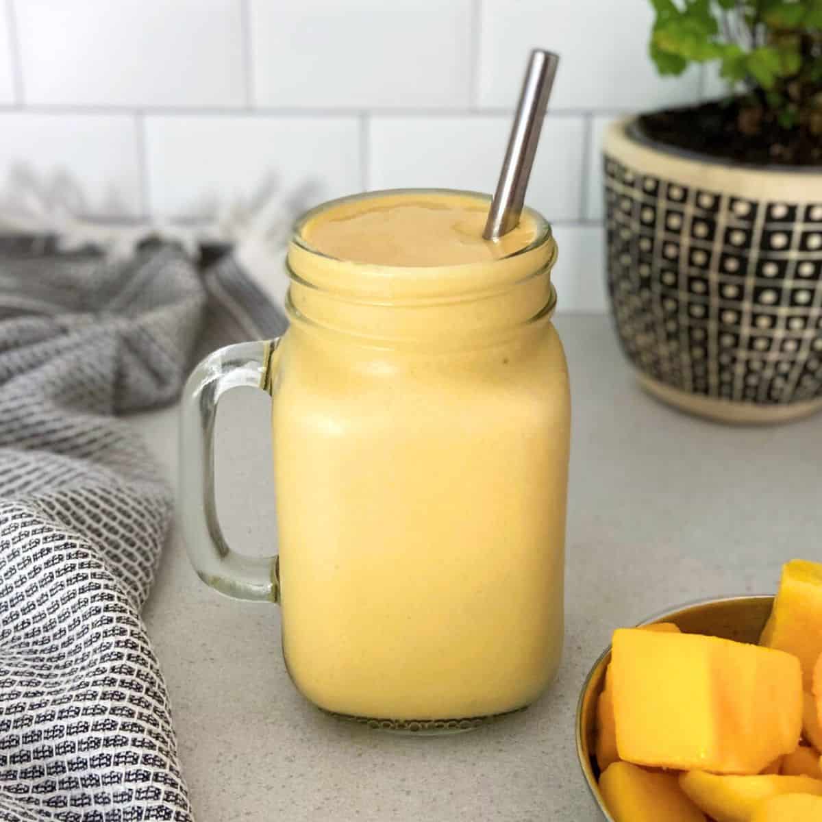 Glass jar filled with a thick and creamy mango protein smoothie, served with a stainless steel straw, a bowl of fresh mango chunks, a patterned cloth napkin, and a decorative plant in the background.