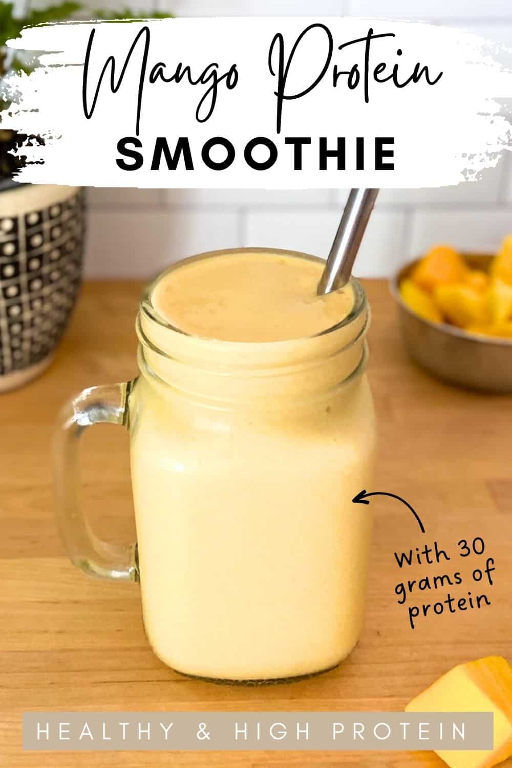 Glass jar filled with a creamy mango protein smoothie on a wooden counter, topped with a metal straw, surrounded by fresh mango pieces, with text overlay highlighting that it has 30 grams of protein.
