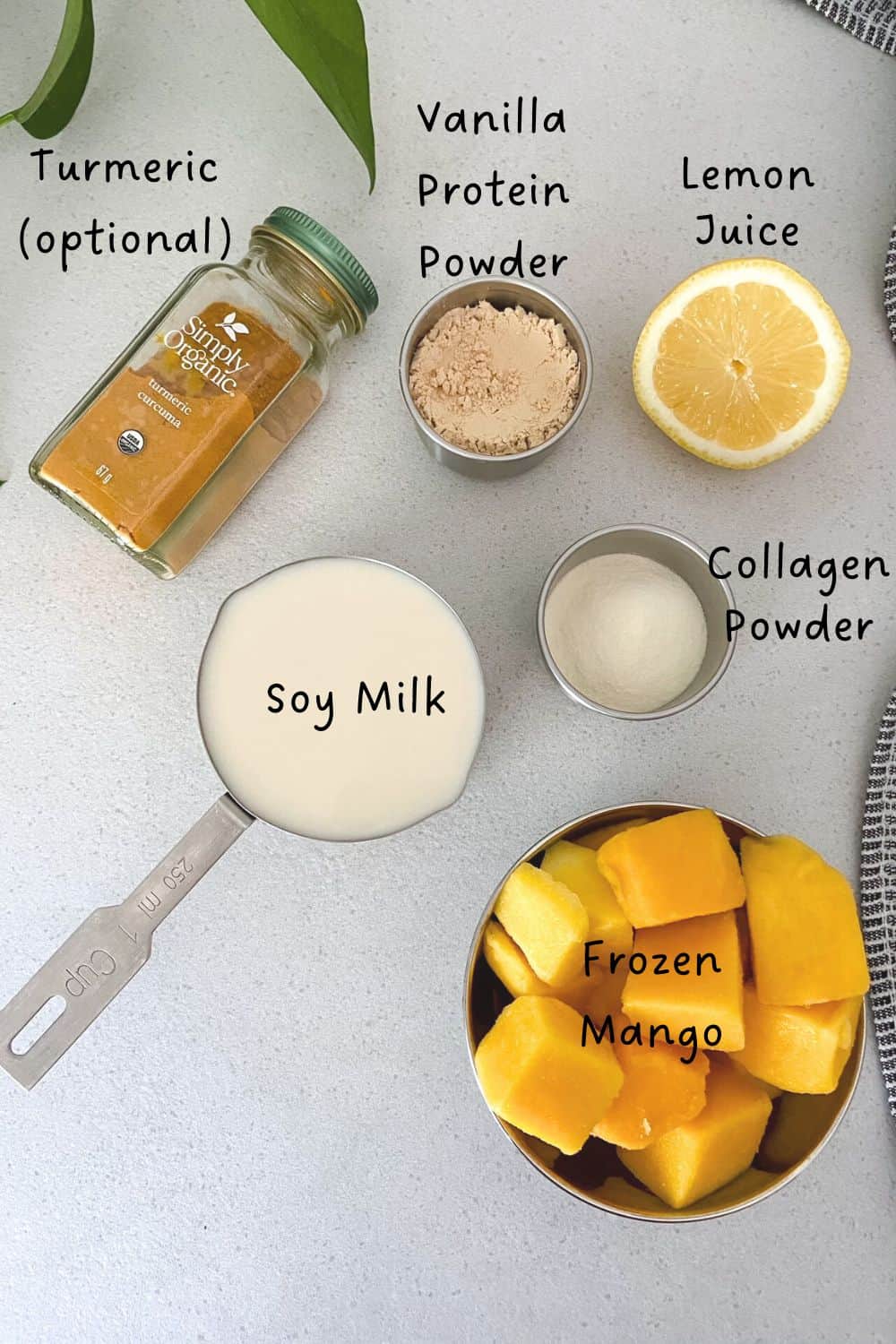 Top-down view of mango protein smoothie ingredients on a white counter, including a bowl of frozen mango cubes, soy milk in a measuring cup, collagen powder, vanilla protein powder, half a lemon, and a jar of turmeric labeled optional.