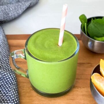 Glass mug filled with a creamy green smoothie with a pink striped straw, served on a wooden board with fresh spinach and frozen mango on the side.