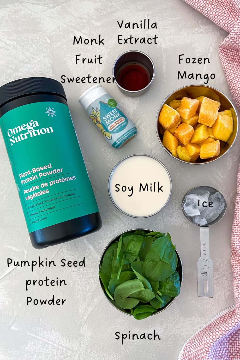 Flat lay of mango spinach protein smoothie ingredients including frozen mango, fresh spinach, soy milk, pumpkin seed protein powder, vanilla extract, monk fruit sweetener, and ice, arranged on a light background with text labels.