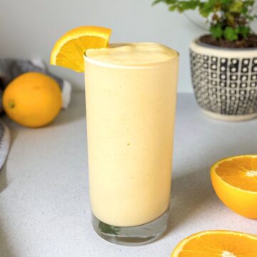 Orange protein smoothie in a tall glass with a creamy thick texture, garnished with a fresh orange slice on the rim, with sliced oranges and a plant in the background.