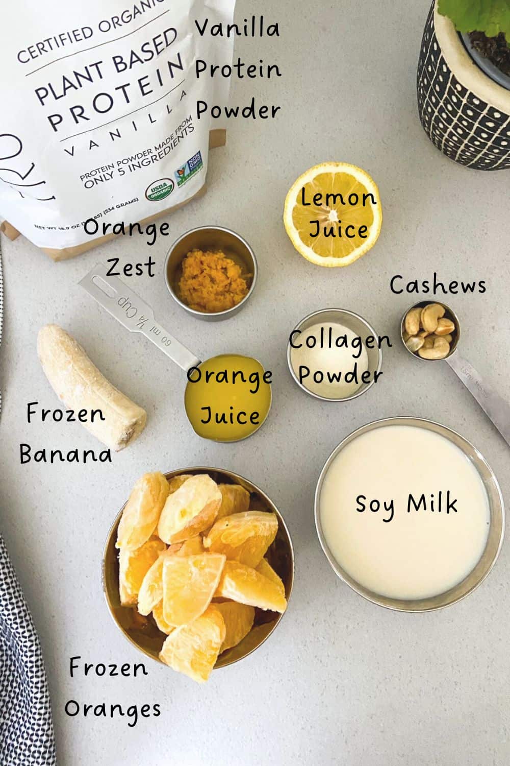 Overhead view of orange protein smoothie ingredients including frozen orange segments, frozen banana, soy milk, cashews, orange juice, lemon juice, orange zest, collagen powder, and vanilla protein powder arranged on a light grey surface.