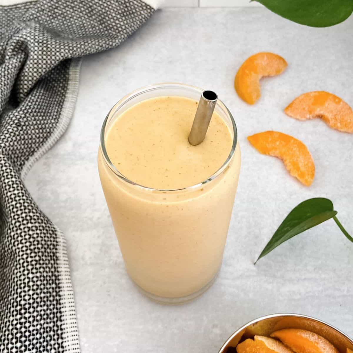 Creamy smoothie in a glass with a metal straw made with frozen peaches, banana, soy milk, and vanilla protein powder. Scattered frozen peaches and a black and white tea towel are beside the smoothie glass.