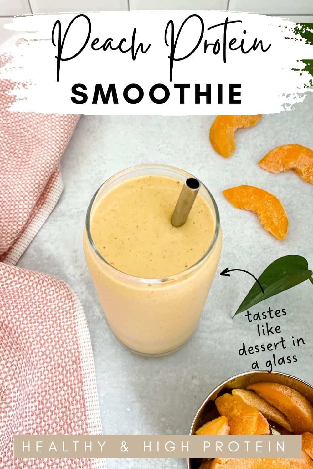 Glass of creamy orange-colored smoothie with a metal straw on a gray surface, next to frozen peach slices, a green leaf, and a pink kitchen towel.