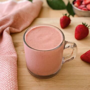 A creamy strawberry protein smoothie in a clear glass mug with a handle on a wooden surface, surrounded by fresh strawberries and a textured pink cloth.