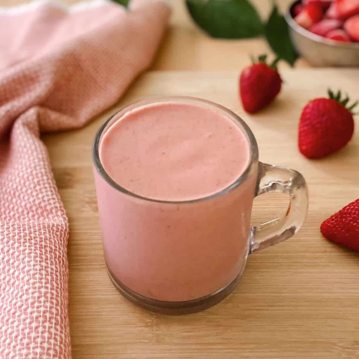 A creamy strawberry protein smoothie in a clear glass mug with a handle on a wooden surface, surrounded by fresh strawberries and a textured pink cloth.