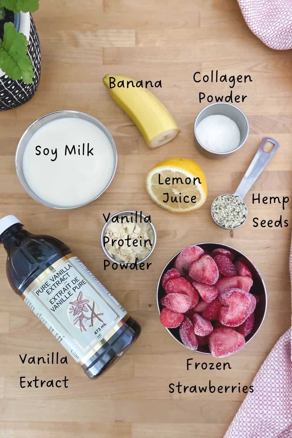 Overhead view of ingredients for a protein strawberry smoothie arranged on a wooden surface, including soy milk, banana, collagen powder, lemon juice, vanilla protein powder, hemp seeds, frozen strawberries, and vanilla extract.