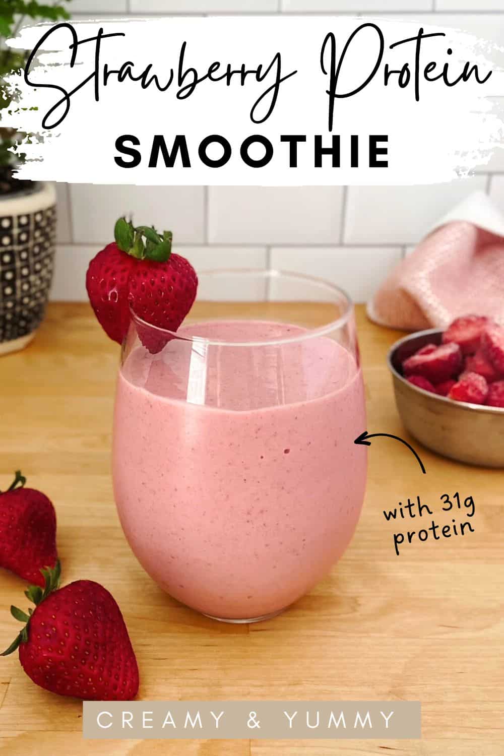 A creamy pink strawberry smoothie in a clear curved glass with a strawberry garnish, set on a wooden surface with fresh strawberries and a bowl of frozen berries in the background.