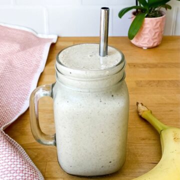 Creamy banana protein smoothie in a glass mason jar with a metal straw on a wooden counter beside a ripe banana and a pink cloth, showing the thick, smooth texture of the drink.