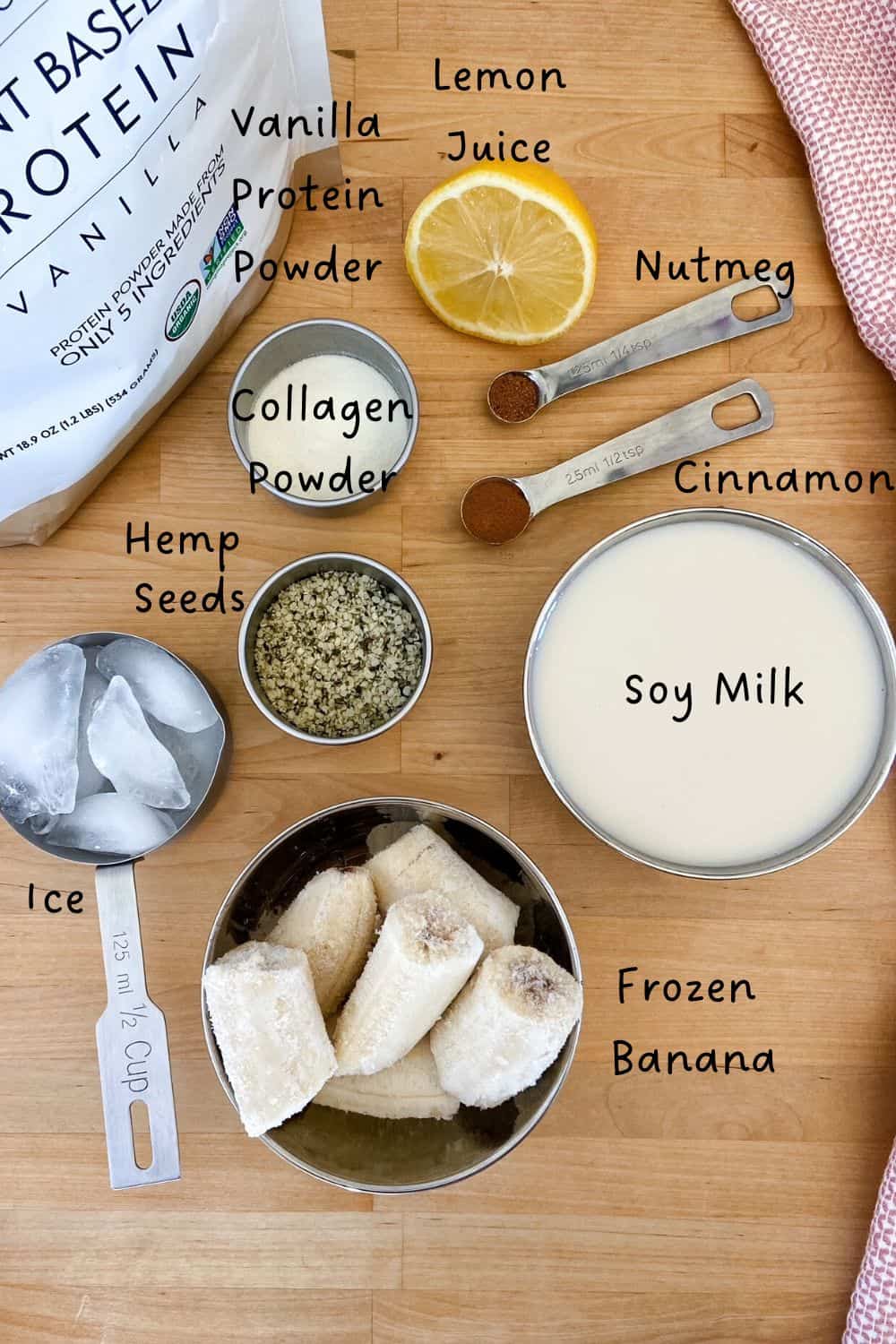 Overhead view of banana protein smoothie ingredients labeled on a wooden counter, including frozen banana, soy milk, vanilla protein powder, collagen peptides, hemp seeds, cinnamon, nutmeg, lemon juice, and ice.
