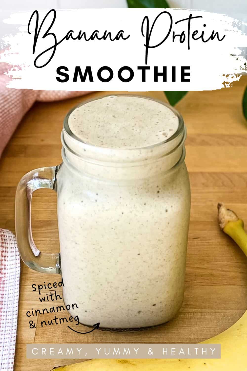 A creamy banana smoothie in a glass mason surrounded by a banana and a pink cloth, with text on the image that says “Banana Protein Smoothie” and “spiced with cinnamon and nutmeg.”