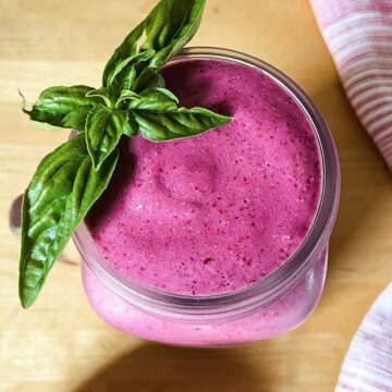 A creamy bright purple blackberry protein smoothie in a glass jar topped with a sprig of fresh basil, sitting on a wooden surface with a pink cloth nearby.