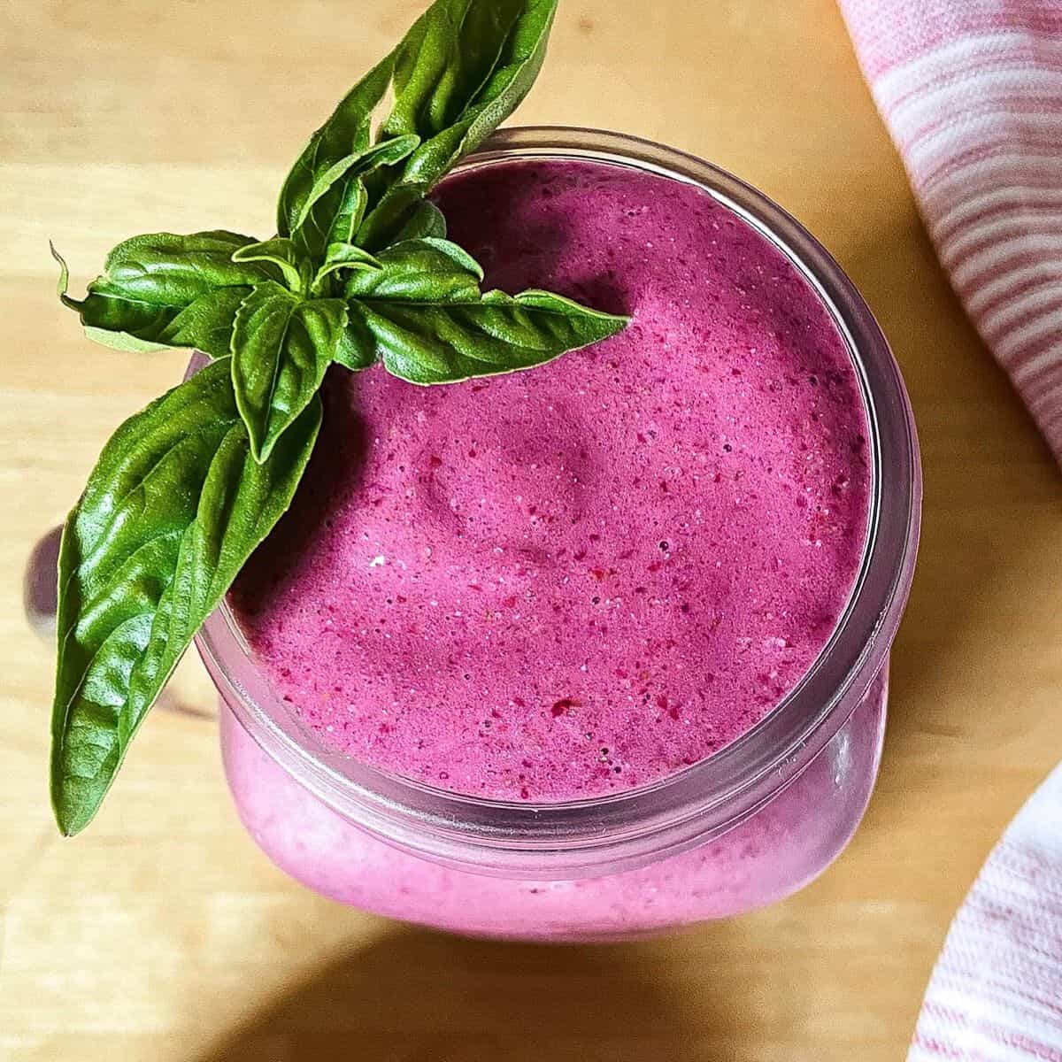 A creamy bright purple blackberry protein smoothie in a glass jar topped with a sprig of fresh basil, sitting on a wooden surface with a pink cloth nearby.