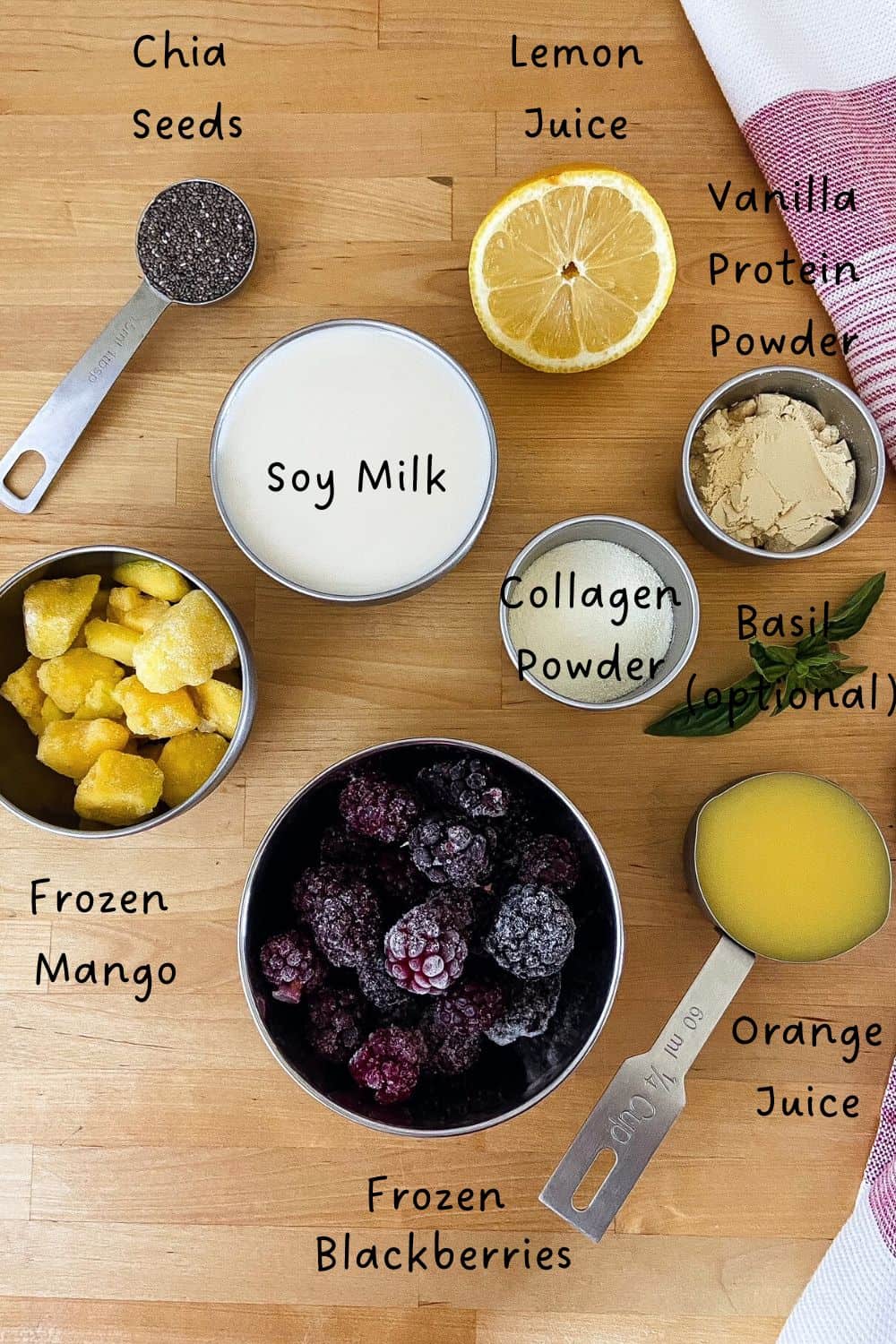 Ingredients for a blackberry protein smoothie laid out on a wooden surface, including frozen blackberries, mango, soy milk, orange juice, lemon juice, chia seeds, protein powder, collagen, and basil leaves.