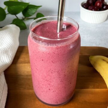 A tall glass filled with a creamy pink cherry banana smoothie with a metal straw, sitting on a wooden board with a banana and a small bowl of cherries in the background.