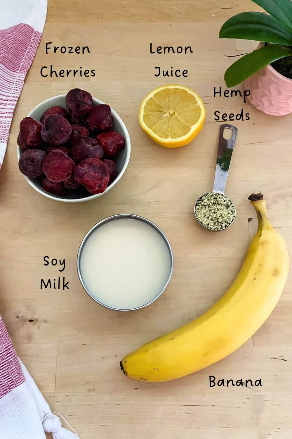 A top down view of the ingredients for a cherry banana smoothie, including a bowl of frozen cherries, a ripe banana, lemon juice, soy milk, and a spoonful of hemp seeds arranged on a light wood surface.
