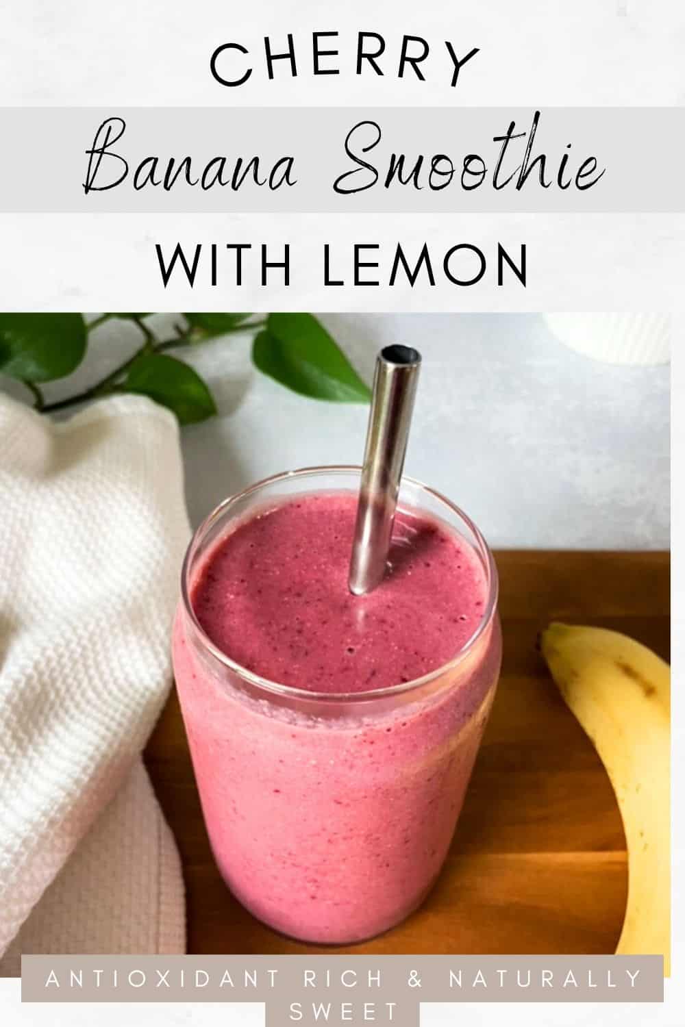 A bright pink smoothie in a clear glass with a metal straw, sitting on a wooden board with a banana nearby and green leaves in the background.