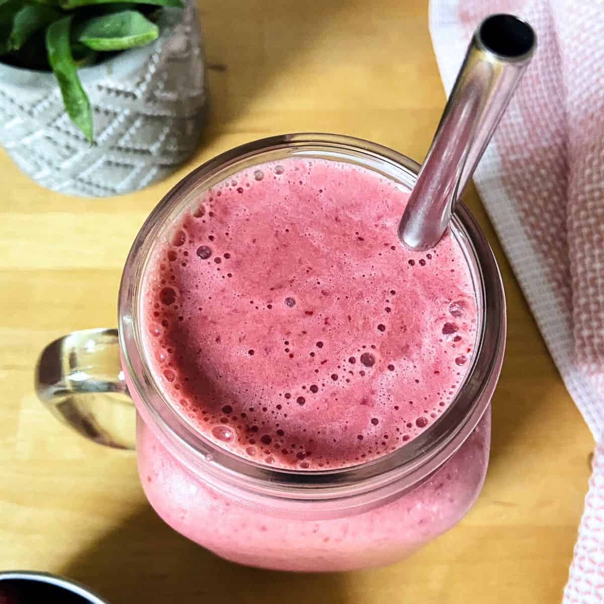 A bright pink smoothie in a glass mason jar with a metal straw on a wooden table, next to a soft pink towel and a small plant in the background.