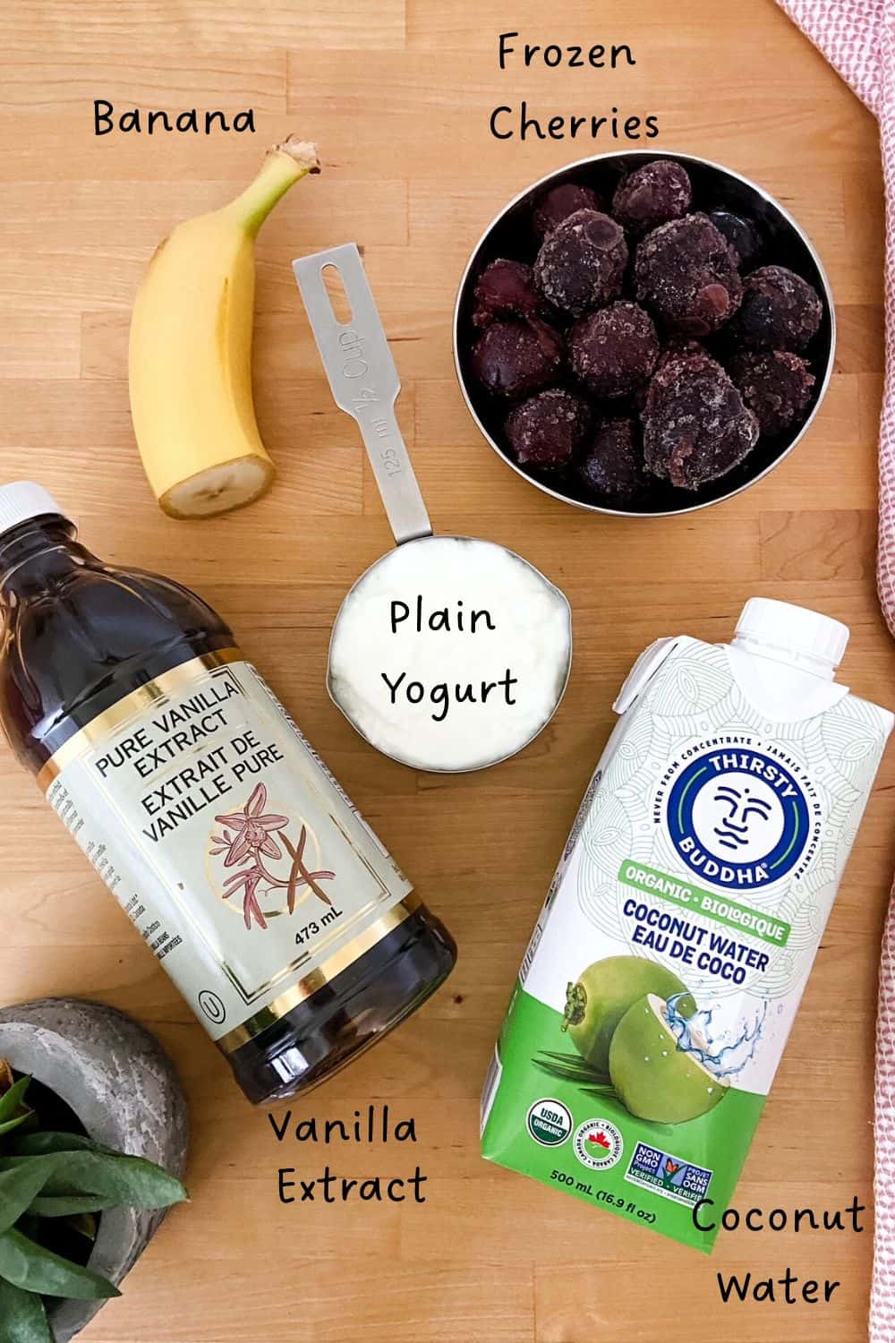 A top down view of frozen cherries, a banana, plain yogurt, vanilla extract, and coconut water arranged on a wood surface.