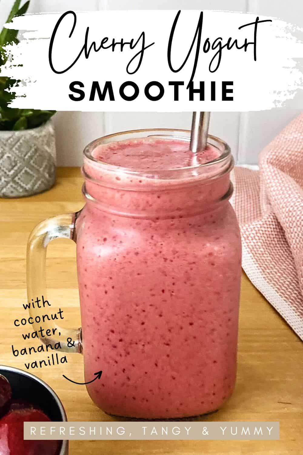 A pink cherry smoothie with yogurt in a mason jar with a metal straw, sitting on a wooden surface with a soft pink cloth in the background.