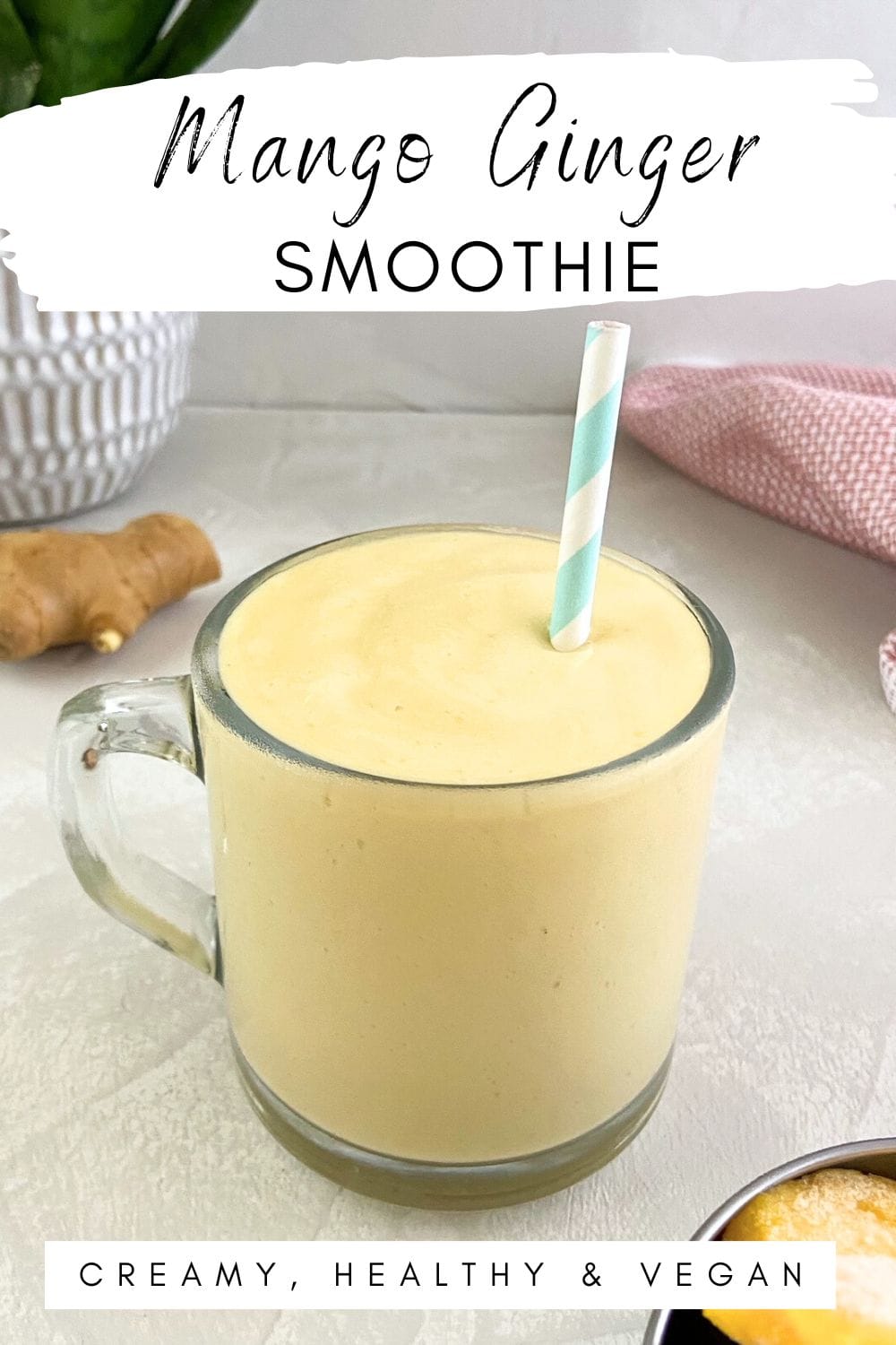 Glass mug of a light yellow smoothie with a striped blue straw, ginger root in the background, pink tea towel, and frozen mango in the corner.