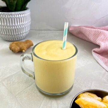Glass mug of creamy mango ginger smoothie with a striped straw, ginger root in the background, a green plant, and a pink tea towel on the side.
