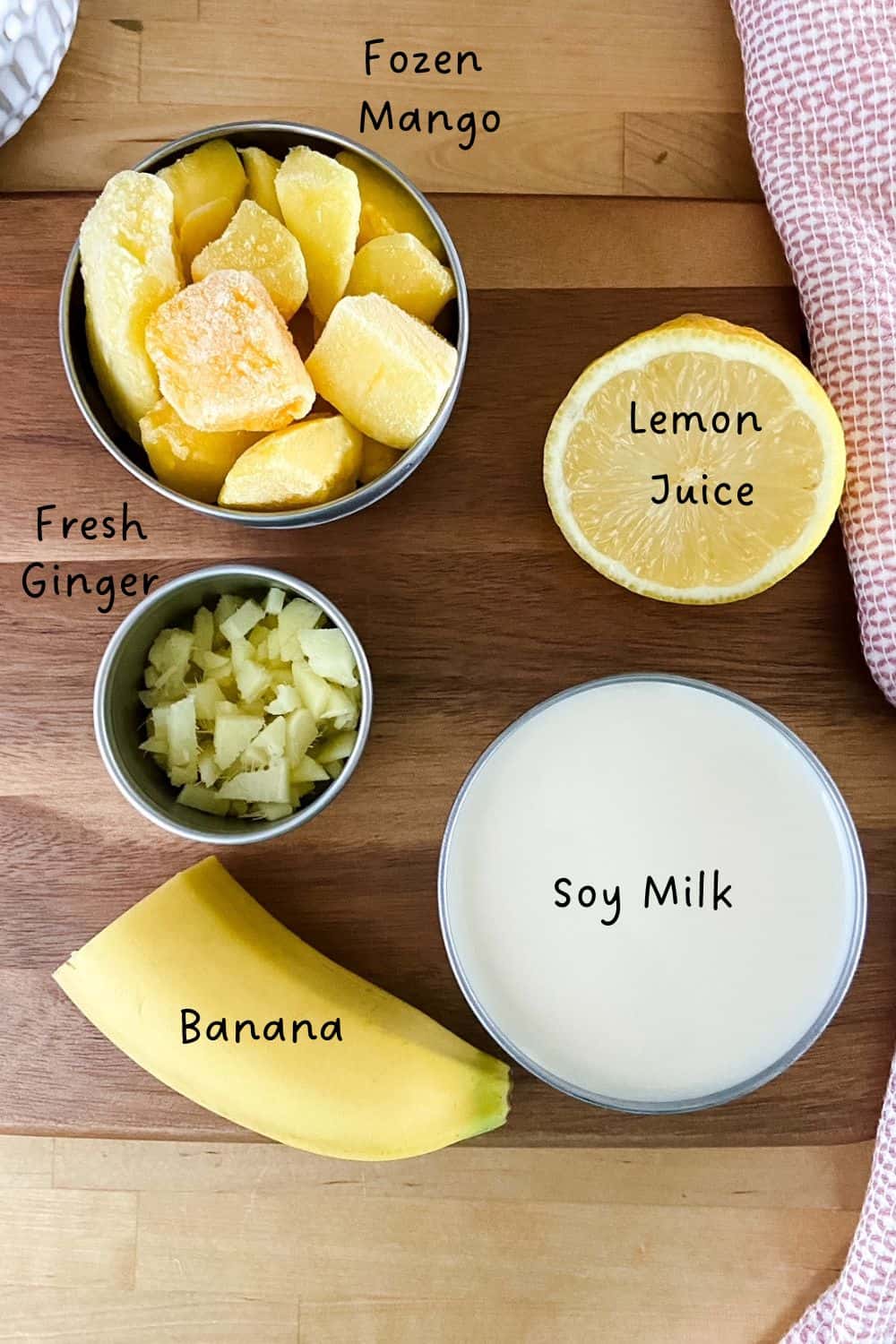 Flat lay of gingermango smoothie ingredients including frozen mango chunks, fresh ginger, soy milk, banana, and lemon on a wooden board with a pink tea towel