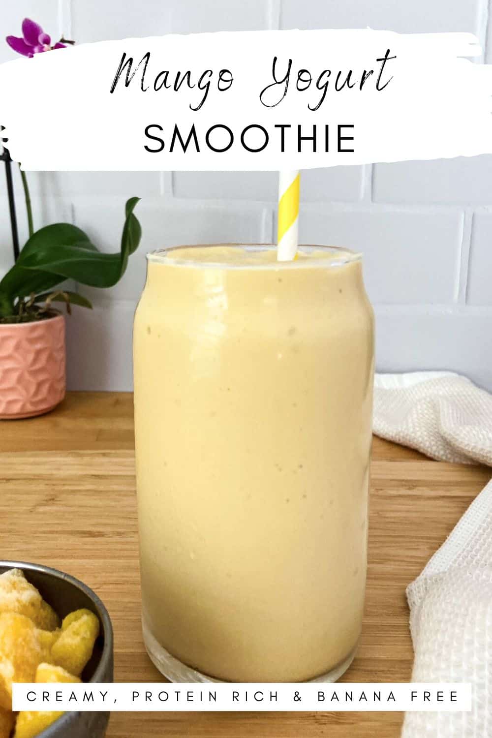 A tall glass of creamy yellow smoothie with a yellow striped straw on a wooden surface, beside a bowl of frozen mango and a pink potted plant in the background.
