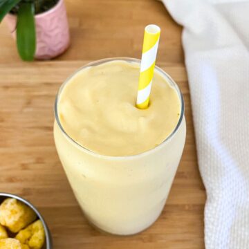 A thick and creamy yellow smoothie served in a clear glass with a yellow-striped straw on a wooden surface, next to a white textured cloth.