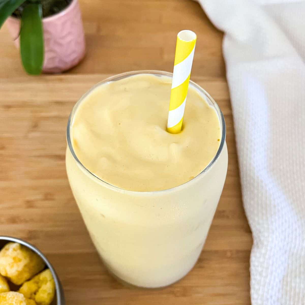 A thick and creamy yellow smoothie served in a clear glass with a yellow-striped straw on a wooden surface, next to a white textured cloth.
