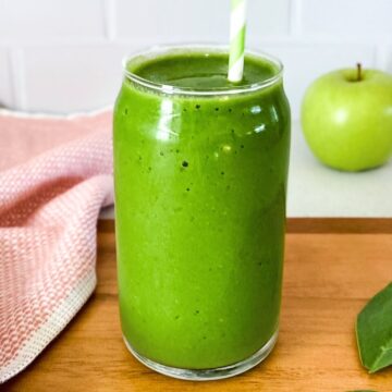 A vibrant green smoothie in a clear glass with a green and white striped straw, sitting on a wooden surface beside a pink towel and a fresh green apple in the background.