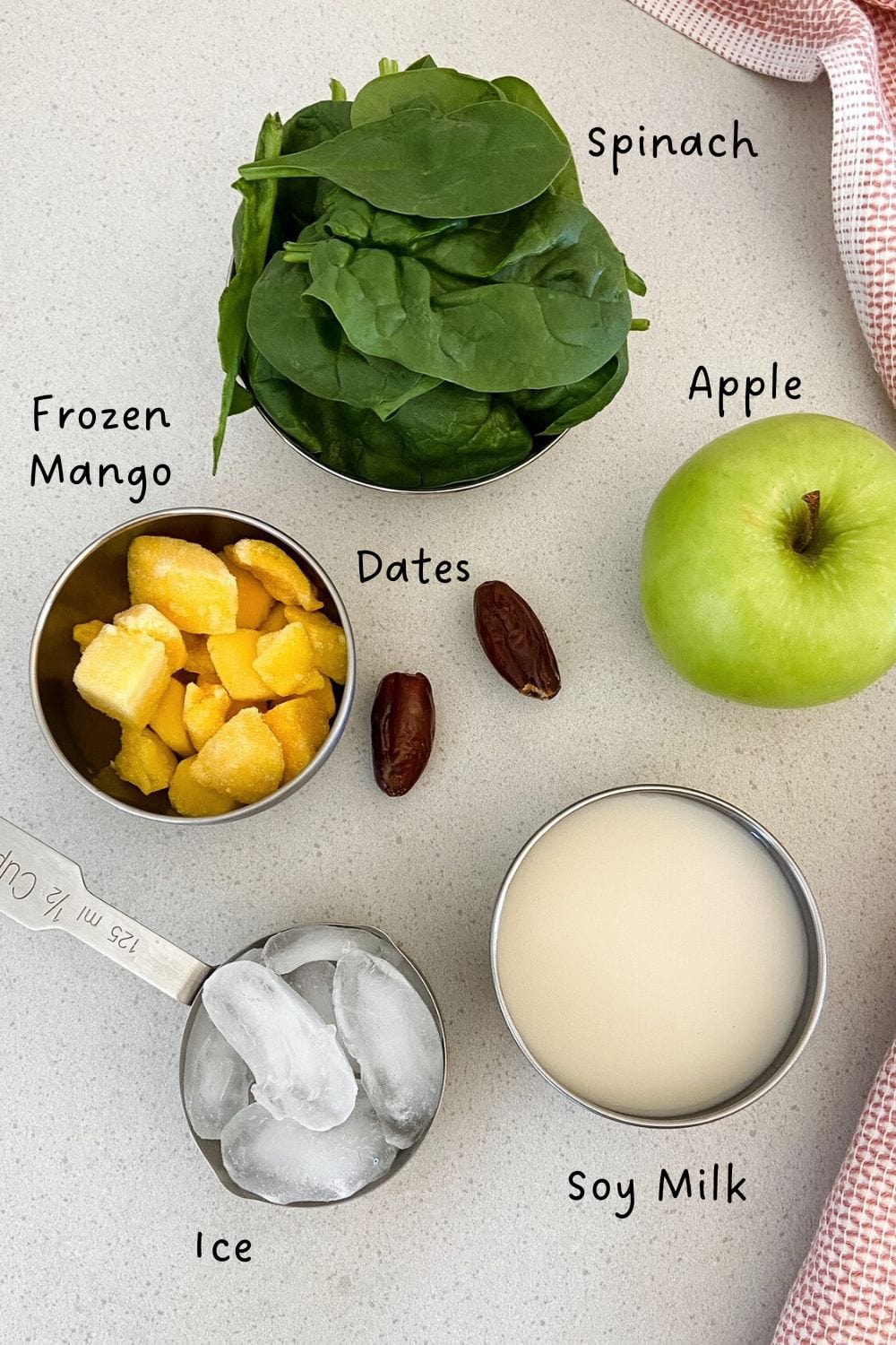 Ingredients for a spinach apple smoothie displayed on a light countertop, including fresh spinach leaves, a green apple, frozen mango chunks, soy milk, dates, and ice, labeled with text.