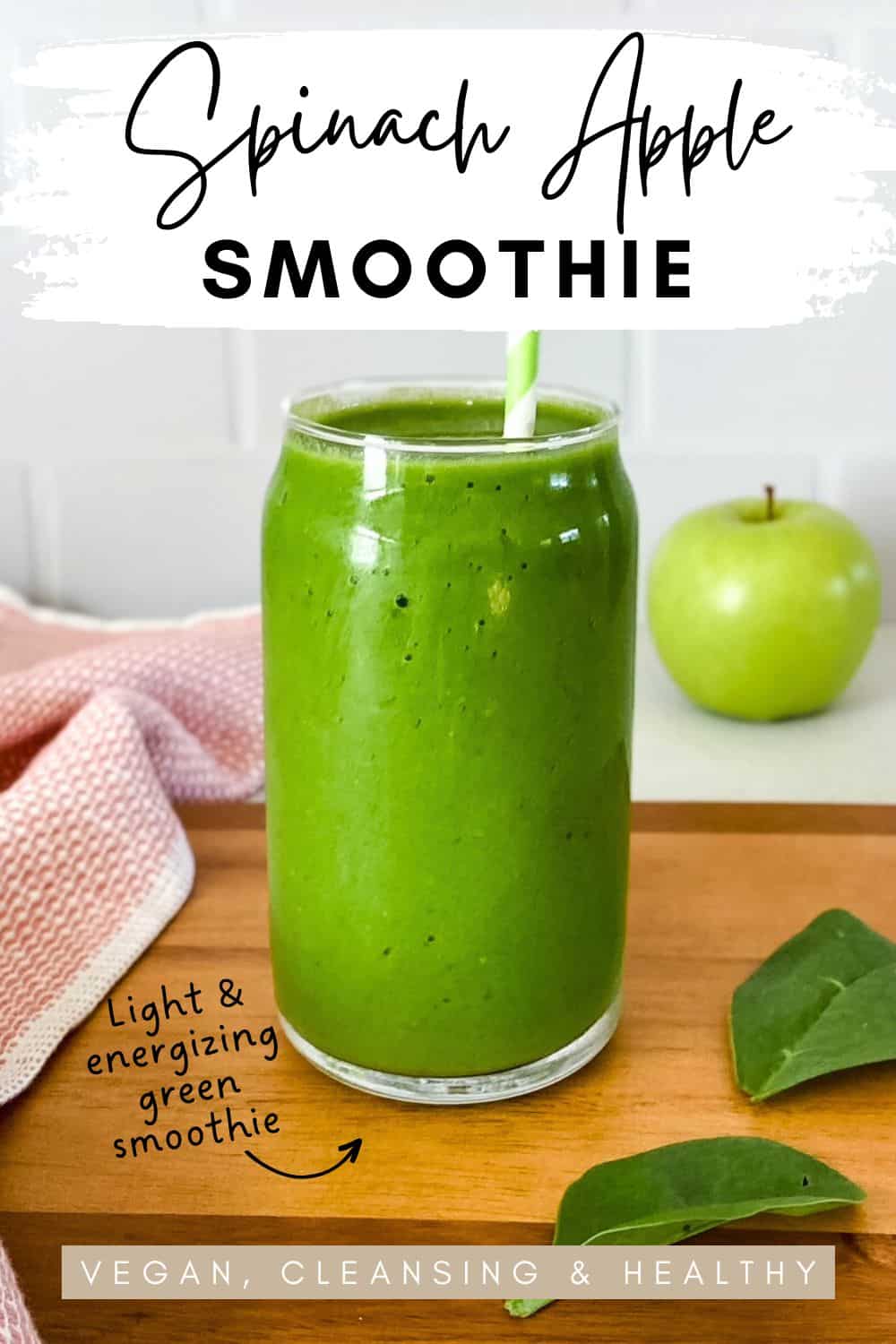 A bright green smoothie in a clear glass with a green and white striped straw, sitting on a wooden surface beside spinach leaves, a green apple, and a pink towel. Text on the image reads spinach apple smoothie vegan, cleansing and healthy.