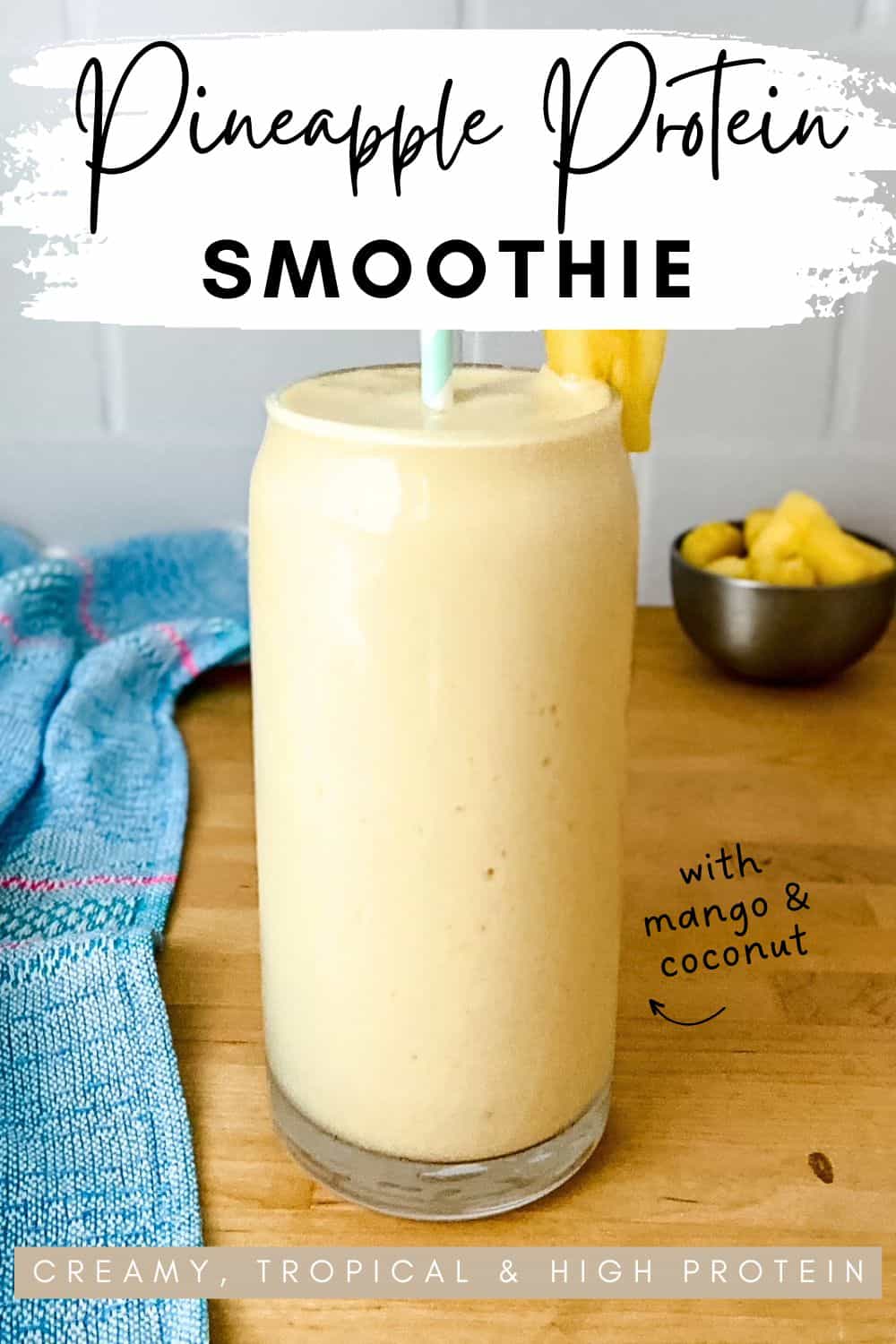 A tall light yellow in a clear glass with a striped straw and a pineapple garnish, shown on a wooden counter with a blue tea towel and a small bowl of pineapple pieces in the background, with text overlay describing the smoothie.