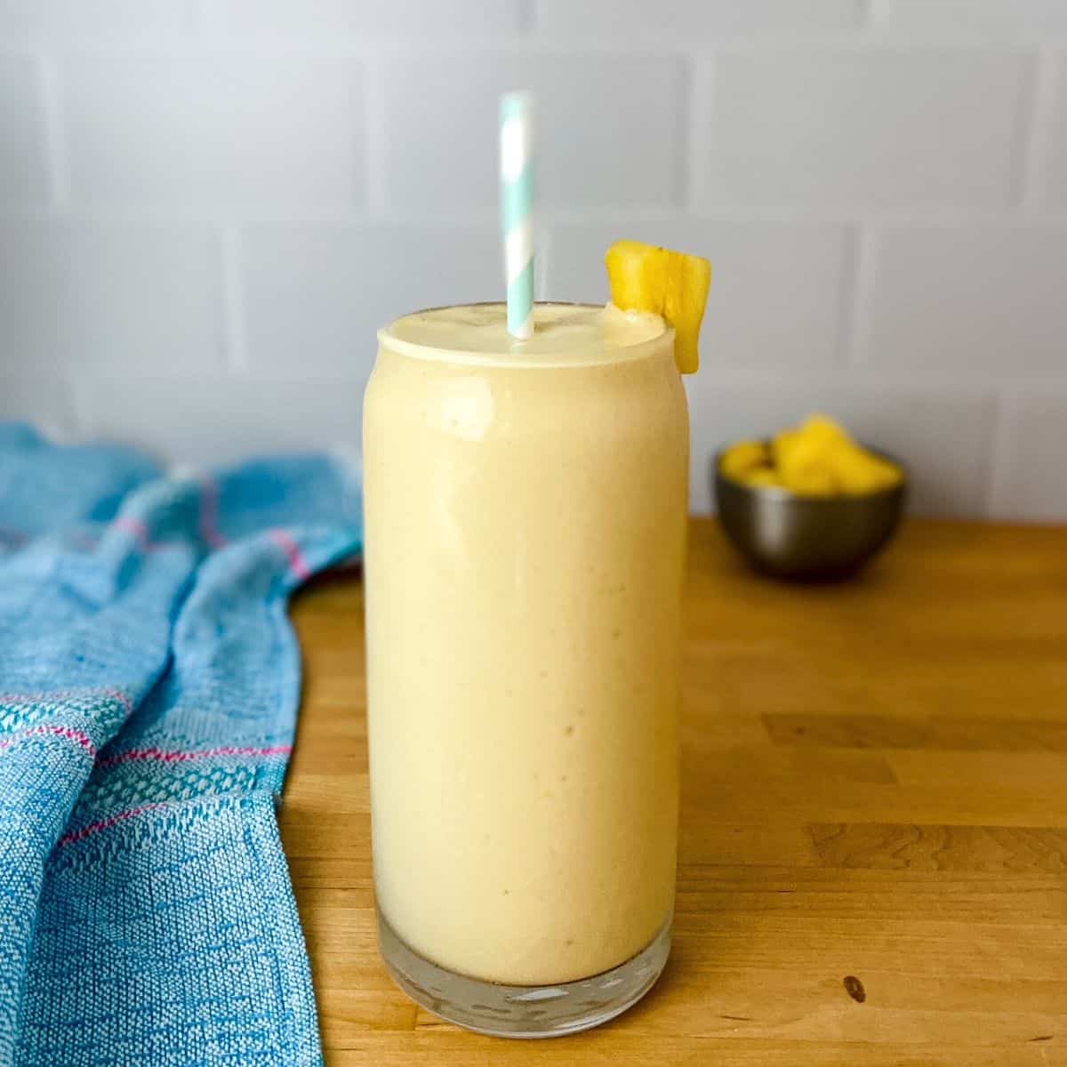 A tall light yellow pineapple protein smoothie in a clear glass with a striped straw and a pineapple garnish, sitting on a wooden counter with a blue tea towel and a small bowl of pineapple pieces in the background.