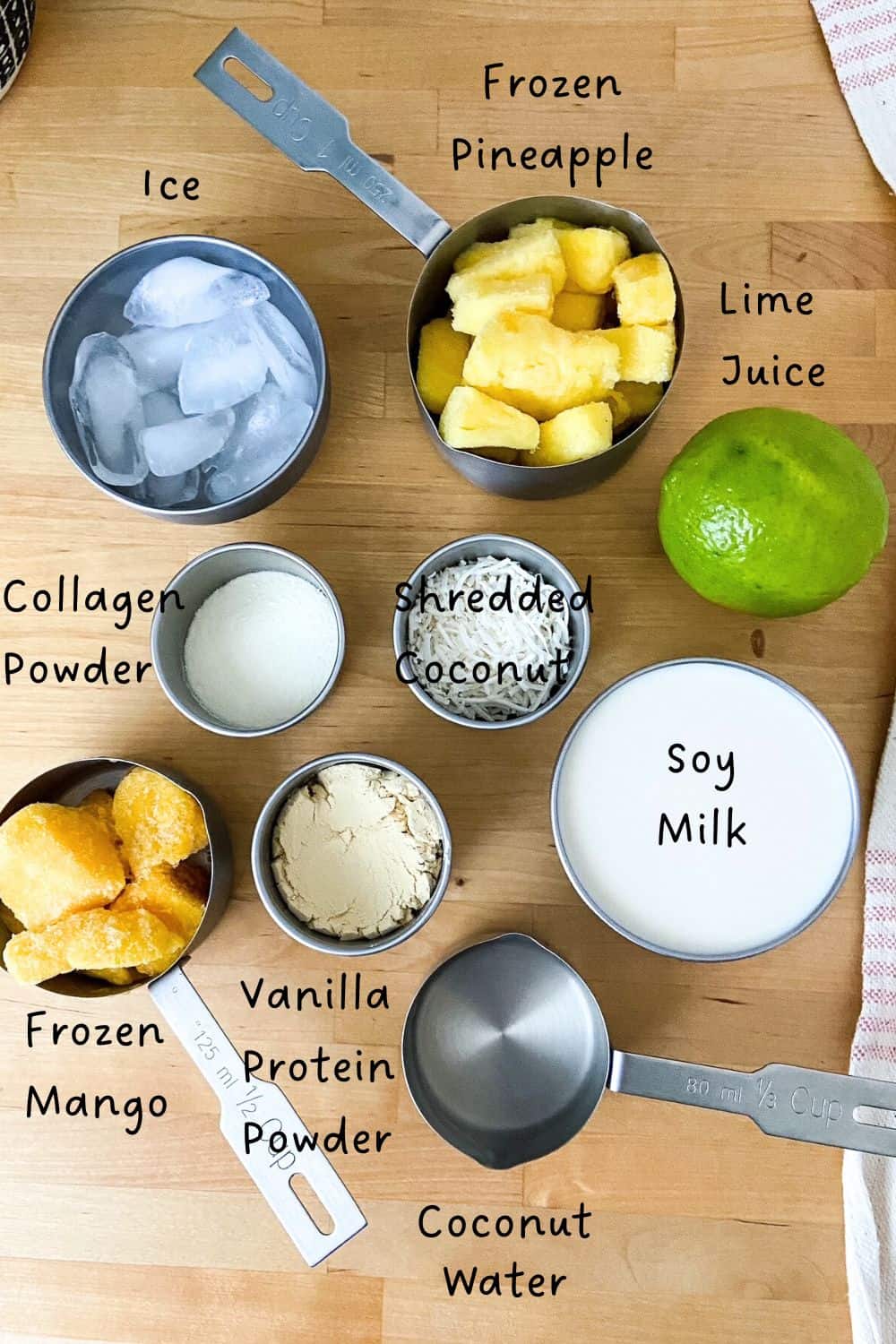 A top view of smoothie ingredients including frozen pineapple, frozen mango, soy milk, lime, shredded coconut, collagen powder, vanilla protein powder, ice, and coconut water arranged on a wooden counter.